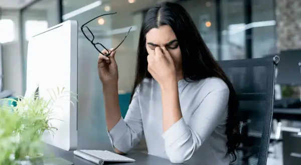 Frau reibt sich die Augen, hält Brille, Computerbildschirm und Tastatur im Büro. Sie braucht eine Pause nach der 20-20-20 Regel.