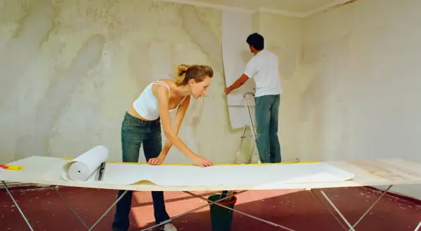 Ein Paar tapeziert eine gestrichene Wand. Die Frau misst Tapete auf einem Tisch, während der Mann sie an der Wand anbringt.
