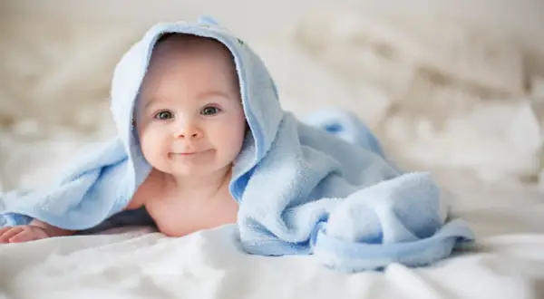Ein lächelndes Baby liegt auf einem Bett, eingewickelt in ein hellblaues Handtuch. Die Frage "wie viel Babykleidung braucht man" beschäftigt viele Eltern.