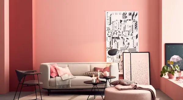 Gemütliches Wohnzimmer mit rosa Wand, bequemer Couch, Kissen und einem Pouf. Ein moderner Sessel und Kunstwerke ergänzen den Raum.