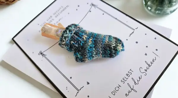 Gestrickte Socke mit Geld als Geschenk. Die Karte fragt "wie viele Socken braucht man?" und ermutigt, sich selbst auf die Socken zu machen.