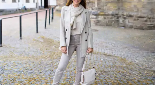 Frau in beige grau farbe Outfit mit Mantel, Schal, Hose und Tasche steht auf Kopfsteinpflaster.