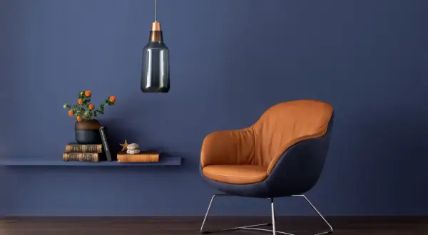 Ein Sessel mit cognacfarbenem Leder und dunkelblauen Akzenten steht vor einer Wand in tiefen blau nuancen. Daneben ein Regal mit Büchern und einer Vase.