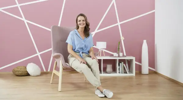 Frau sitzt in einem Sessel vor einer Wand mit geometrischem **Wanddesign streichen**. Ein Beistelltisch mit Deko steht daneben.