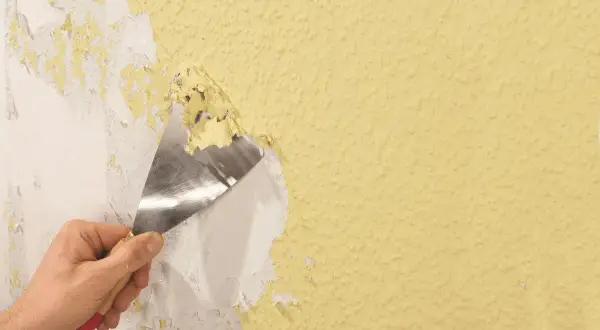 Hand schabt mit Spachtel alte Tapetenreste von gelber Wand.