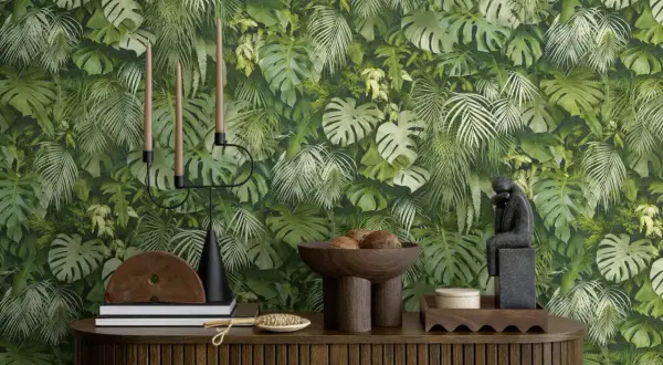 Dschungel-Tapete mit Monstera und Farnen. Ein Kerzenständer, Bücher und eine Skulptur stehen auf einem Holzmöbel. Der Unterschied Vlies und Papiertapete wird hier nicht gezeigt.