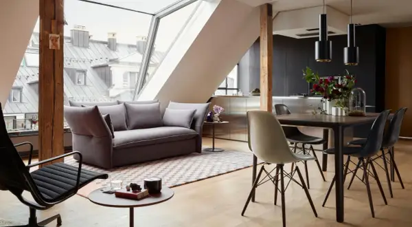Modernes Wohnzimmer mit Dachschräge, wo sich Sofa und Esstisch kombinieren. Holzbalken und große Fenster schaffen ein luftiges Ambiente.