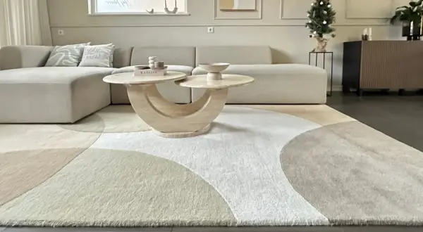 Ein Wohnzimmer mit einem beigen Ecksofa, einem runden Couchtisch und einem modernen Teppich im geometrischen Design. Dieser Teppich-Trend bringt Ruhe und Stil in jeden Raum.