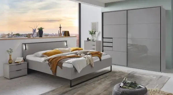 Modernes Schlafzimmer mit grauem Bett, Nachttischen und einem großen Kleiderschrank. Ein Fenster gibt den Blick auf eine Stadt bei Sonnenuntergang frei.