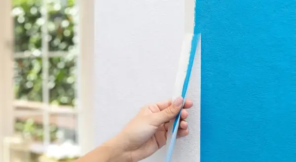 Hand zieht blauen Malerkrepp von weißer Wand ab. Das ist ein wichtiger Schritt, wenn man wissen will, was man zum Streichen braucht.