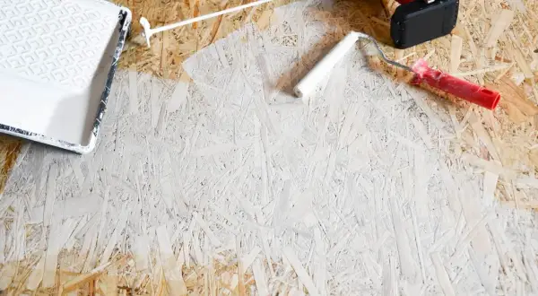 OSB-Platten streichen: Ein weißer Farbroller und eine Farbwanne liegen bereit. Ein Akkuschrauber und Handschuhe deuten auf ein DIY-Projekt hin.