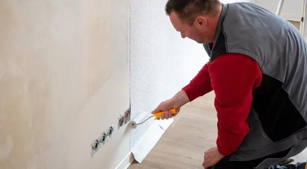 Mann tapeziert eine Wand. Der Kleisterroller wird auf den Putz aufgetragen, bevor die Tapete angebracht wird.