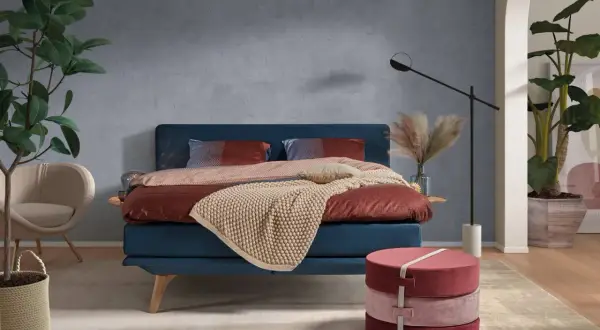 Ein blaues Bett mit rustikaler, grauer Wandfarbe im Hintergrund. Dekorative Kissen und eine warme Decke laden zum Entspannen ein.