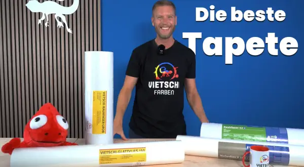 Mann vergleicht Malervlies vs Rauhfaser Kosten. Mehrere Rollen Tapeten liegen auf einem Tisch, ein Plüschtier schaut hervor.