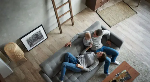 Familie entspannt auf Sofa, ein Baby auf dem Arm. Minimalismus Lebensstil in einem aufgeräumten, hellen Wohnzimmer mit Holzboden.