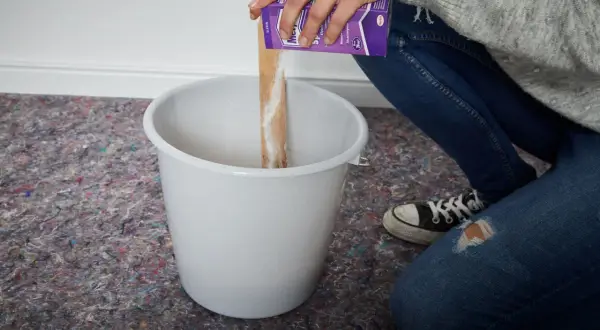 Hände schütten Pulver aus einer Packung in einen Eimer, um Tapetenkleister aufzutragen.
