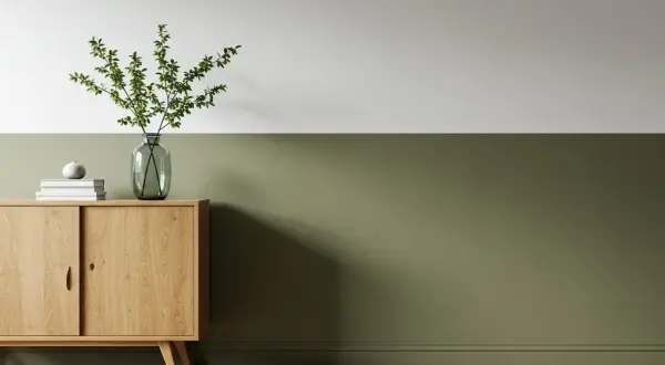 Ein Sideboard aus Holz mit Büchern und einer Vase mit Zweigen steht vor einer zweifarbig gestrichenen Wand. Die untere Hälfte der Wand ist in einem erdigen Grün gehalten, die obere Hälfte in Weiß.