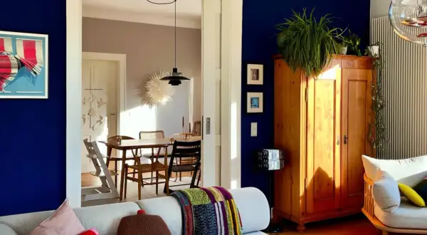 Ein Wohnzimmer mit einer tiefblauen Wand, einem Holzkasten, Pflanzen und einem gemütlichen Sofa. Durch eine Tür ist ein Esszimmer mit Holztisch und Stühlen zu sehen.