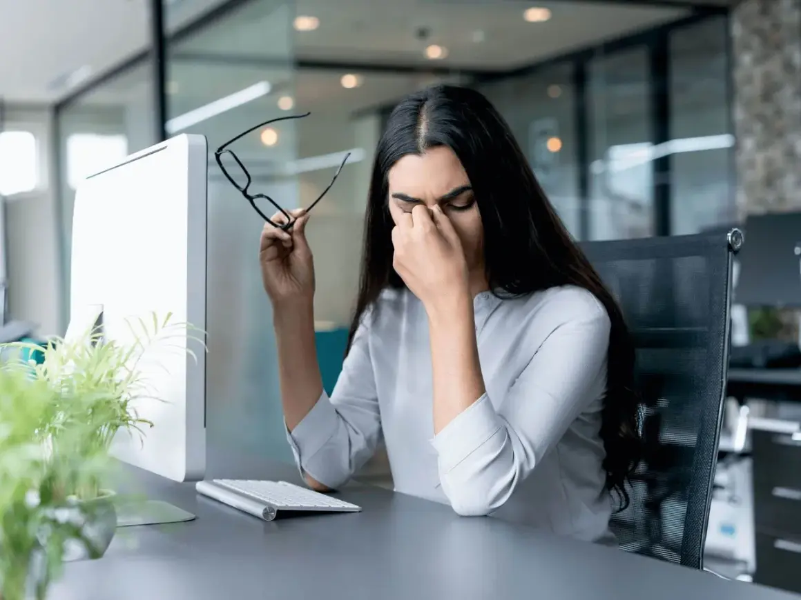 Frau reibt sich die Augen, hält Brille, Computerbildschirm und Tastatur im Büro. Sie braucht eine Pause nach der 20-20-20 Regel.