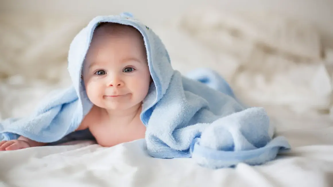 Ein lächelndes Baby liegt auf einem Bett, eingewickelt in ein hellblaues Handtuch. Die Frage "wie viel Babykleidung braucht man" beschäftigt viele Eltern.