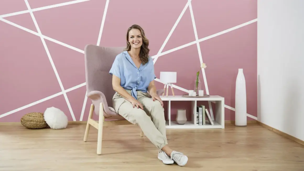 Frau sitzt in einem Sessel vor einer Wand mit geometrischem **Wanddesign streichen**. Ein Beistelltisch mit Deko steht daneben.