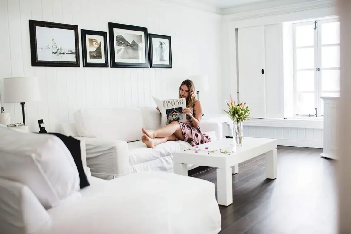 Frau liest Magazin auf weißem Sofa, umgeben von minimalistisch eingerichteten Räumen. Helle Farben und klare Linien schaffen eine ruhige Atmosphäre.