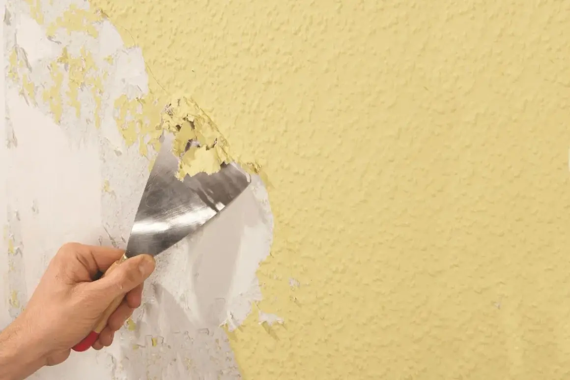 Hand schabt mit Spachtel alte Tapetenreste von gelber Wand.
