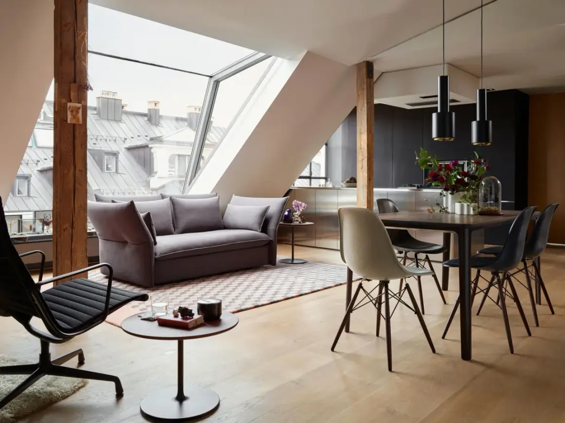 Modernes Wohnzimmer mit Dachschräge, wo sich Sofa und Esstisch kombinieren. Holzbalken und große Fenster schaffen ein luftiges Ambiente.