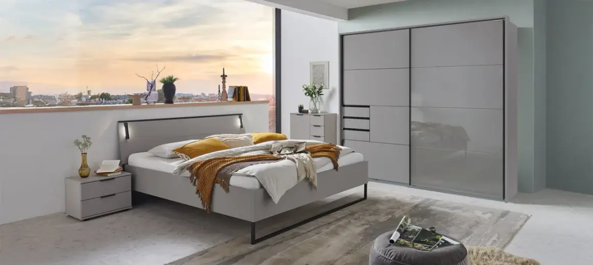Modernes Schlafzimmer mit grauem Bett, Nachttischen und einem großen Kleiderschrank. Ein Fenster gibt den Blick auf eine Stadt bei Sonnenuntergang frei.