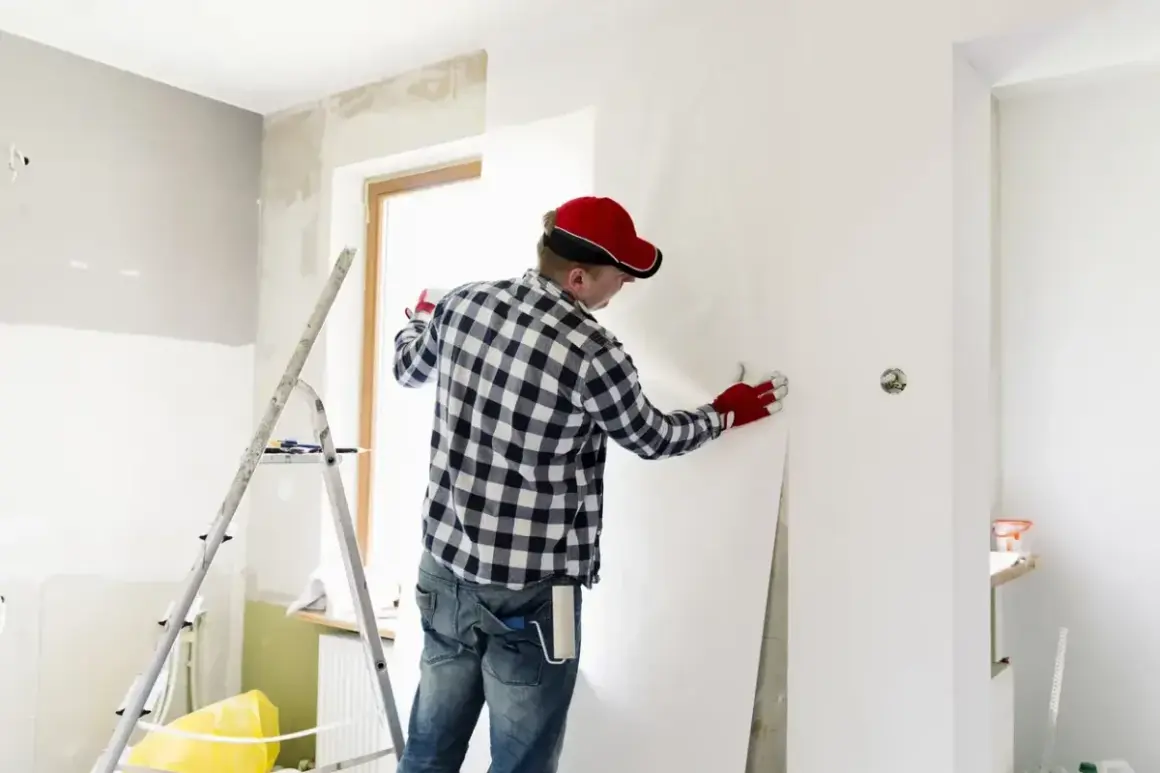 Mann mit roter Kappe tapeziert Wand. Verschiedene Raufasertapeten Arten sind für Renovierungen beliebt.