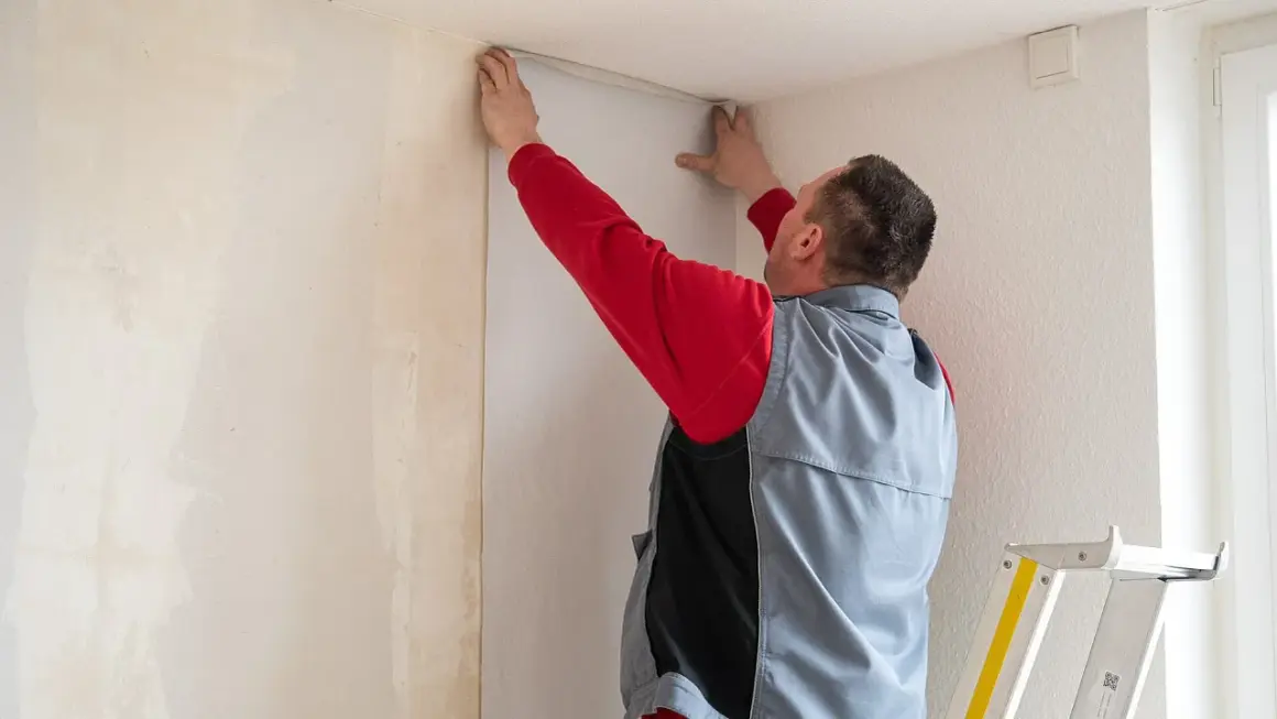 Mann tapeziert eine Wand in einer Wohnung. Er hält eine Tapetenbahn an die Decke, um sie anzubringen.