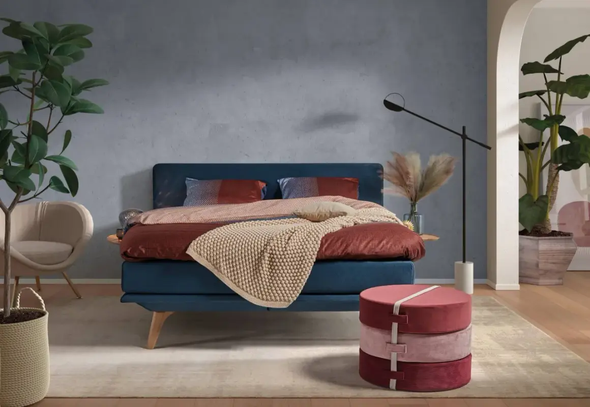 Ein blaues Bett mit rustikaler, grauer Wandfarbe im Hintergrund. Dekorative Kissen und eine warme Decke laden zum Entspannen ein.