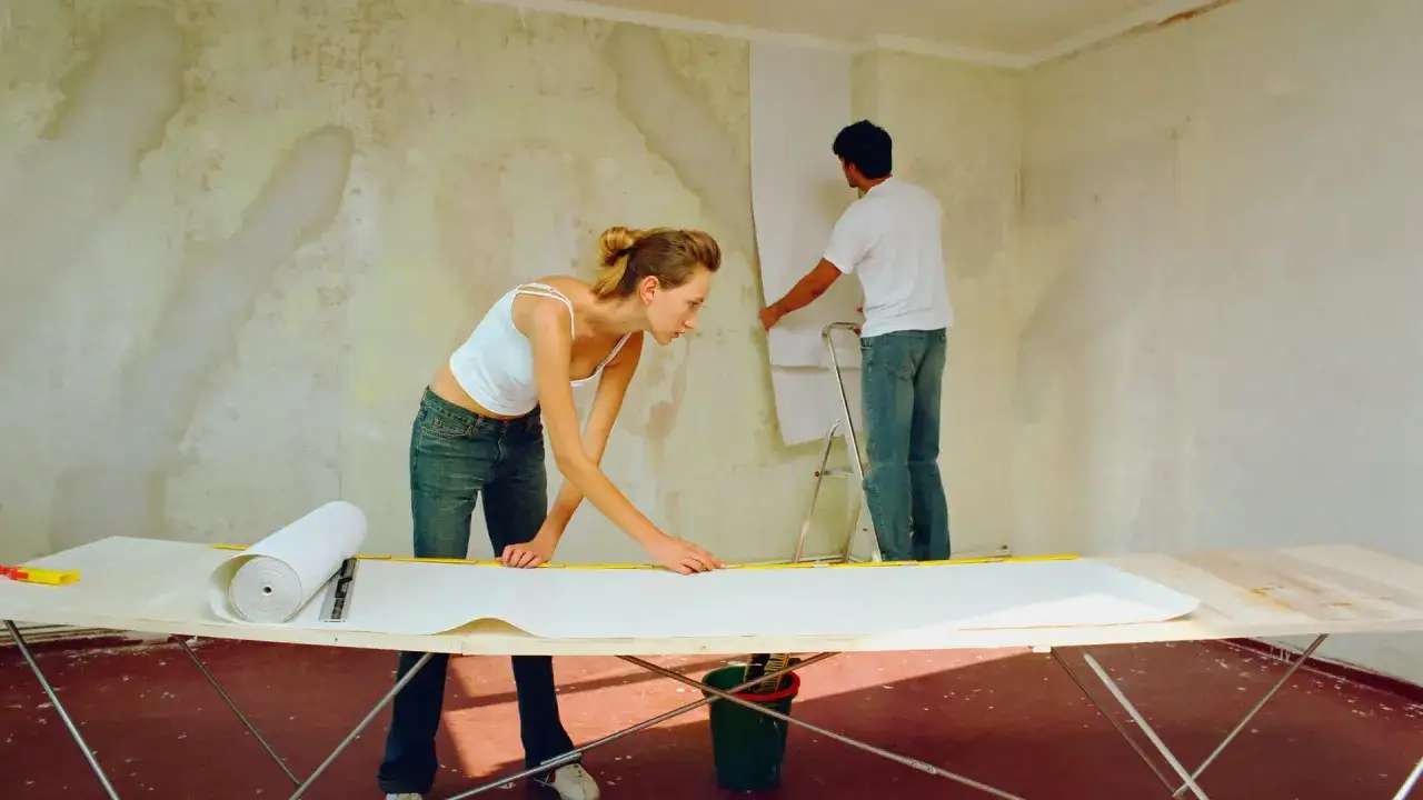 Ein Paar tapeziert eine Wand. Die Frau misst die Tapete auf einem Tisch, während der Mann sie an der gestrichenen Wand anbringt.