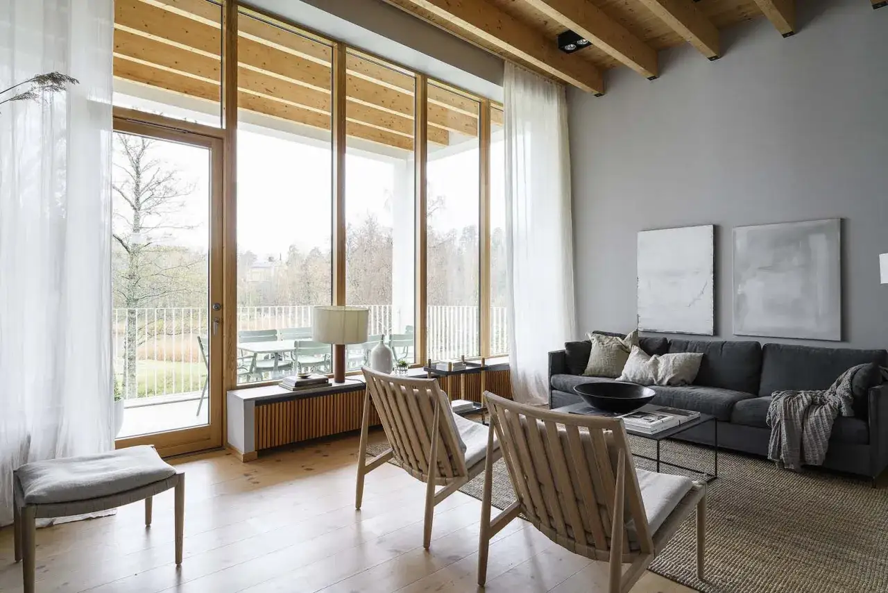 Gemütliches Wohnzimmer mit warmes grau farbpalette, Holzbalken und großen Fenstern, die auf eine Terrasse mit Blick ins Grüne führen.