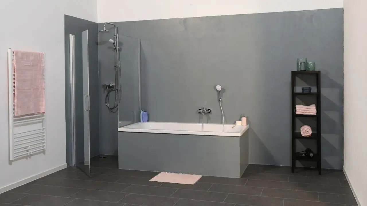 Modernes Bad mit grauen W&auml;nden, die wie Betonfarbe aussehen. Die Badewanne ist ebenfalls in Betonoptik gehalten. Handt&uuml;cher und Dekoartikel sind auf einem schwarzen Regal platziert.