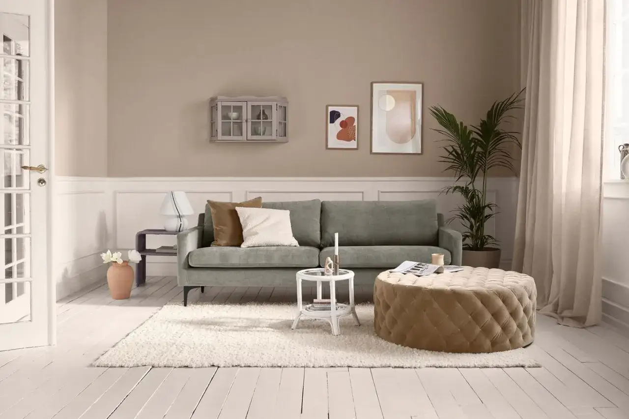 Gemütliches Wohnzimmer mit beigem Teppich, grauem Sofa und modernen Wandfarben. Ein Hocker und Kunstwerke runden das Bild ab.