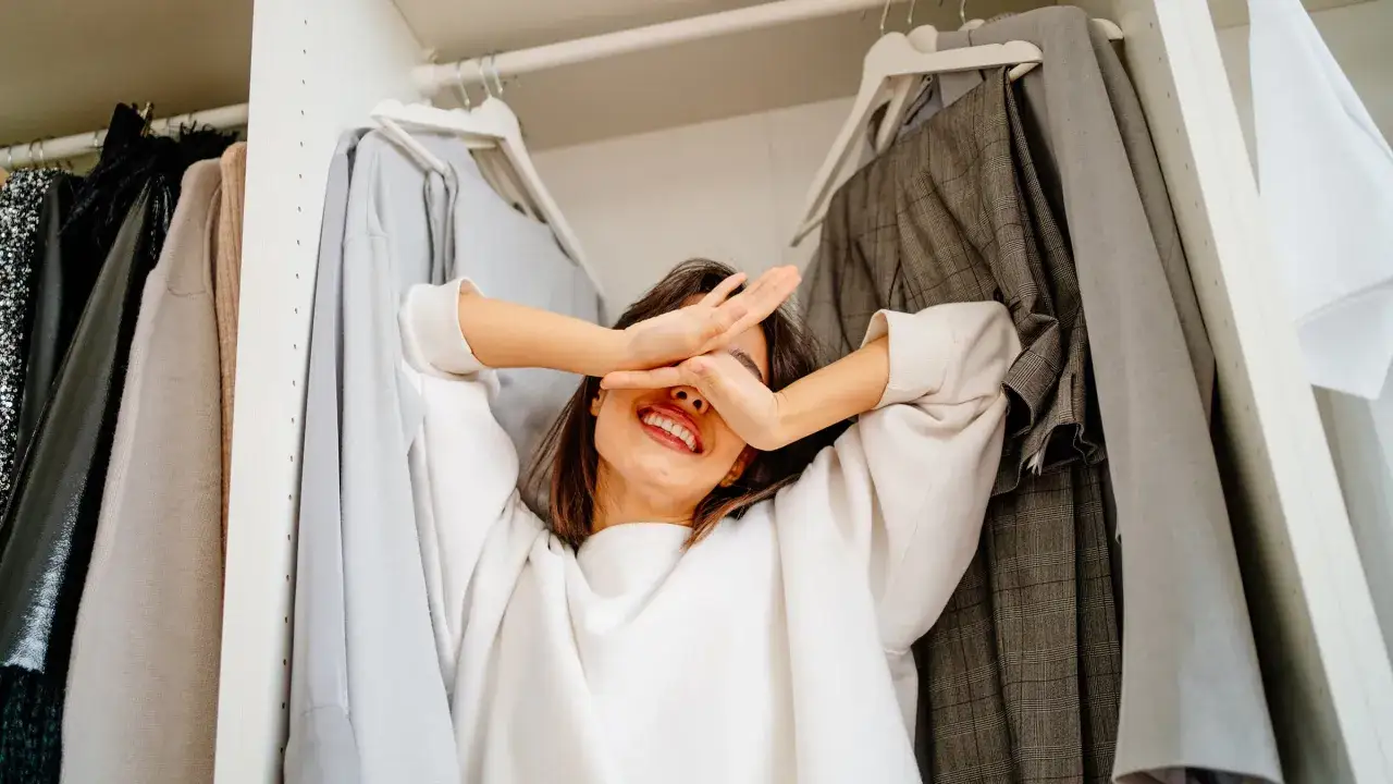 Frau versteckt sich lachend im Kleiderschrank, umgeben von Kleidung. Zeit, Kleidung zu reduzieren und Platz zu schaffen!