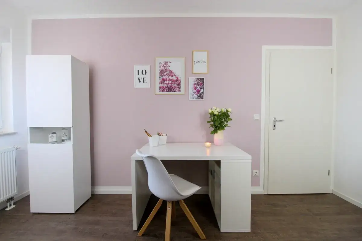 Schreibtisch mit Blumen und Kerze vor einer zartrosa Wand, die nicht bis zur Decke gestrichen ist. Ein weißer Schrank und eine Tür sind ebenfalls zu sehen.
