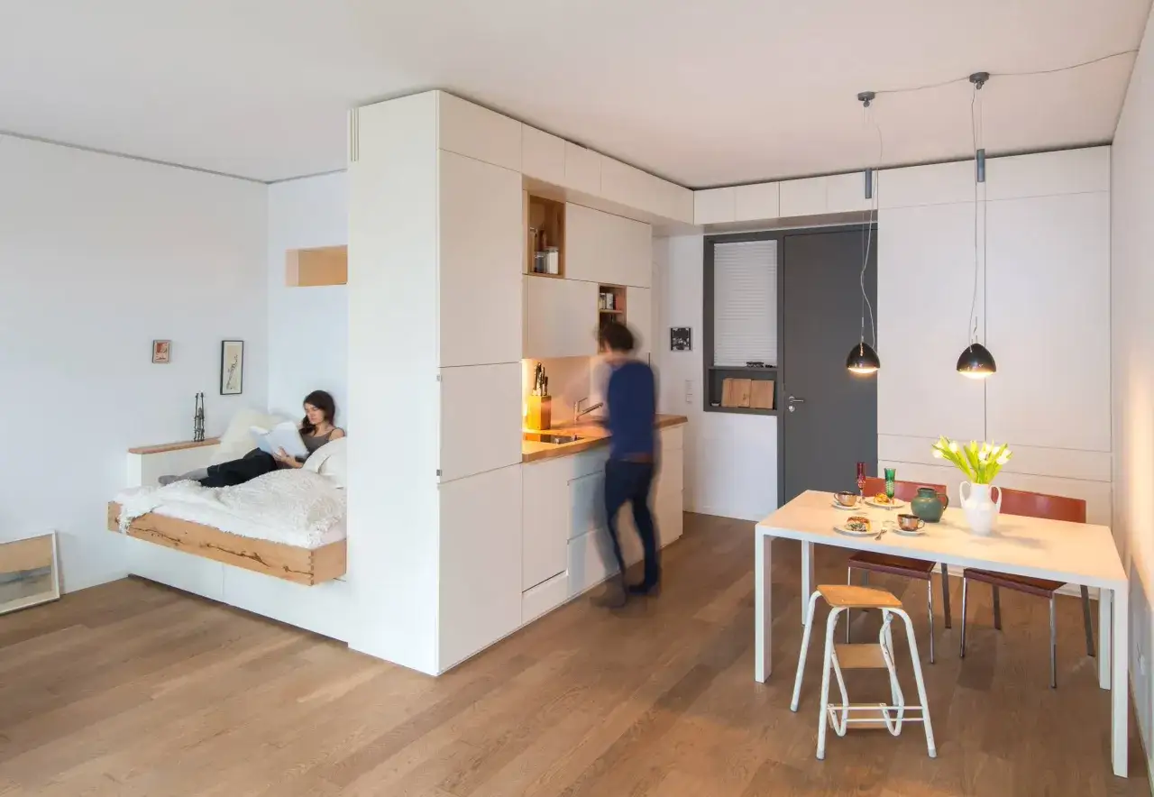 Gem&uuml;tliche Wohnung mit Bettnische, K&uuml;che und Esstisch. Eine Frau liest im Bett, w&auml;hrend jemand in der K&uuml;che besch&auml;ftigt ist. Zeit, das handy weglegen und genie&szlig;en.