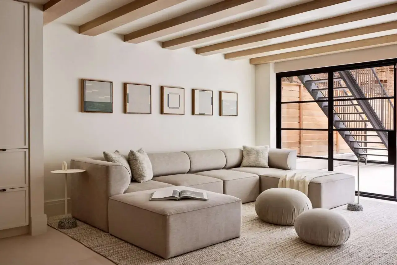 Gemütliches Wohnzimmer mit beiger Couch, Holzbalken und Kunst an der Wand. Die Wandfarbe cosy kombinieren ist hier perfekt umgesetzt.