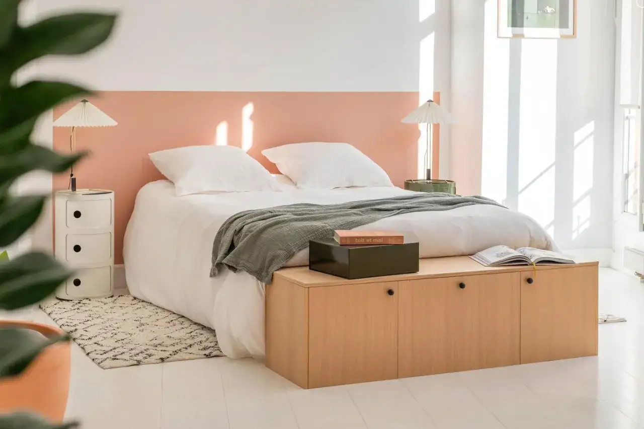 Schlafzimmer mit wei&szlig;em Bett, Laken und gr&uuml;ner Decke. Die Wand nur teilweise streichen in zartem Rosa. Holzbank mit B&uuml;chern.