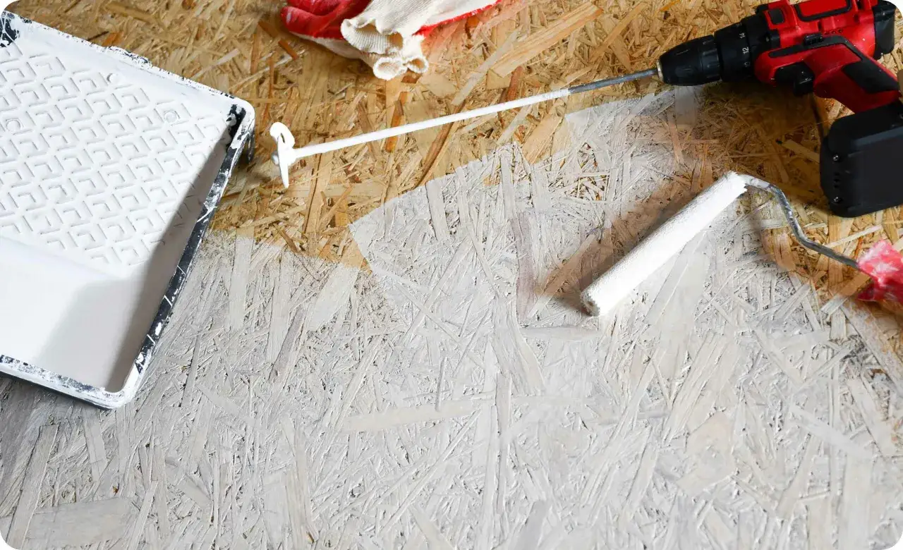 Arbeiten an OSB-Platten: Ein weißer Farbroller und eine Farbwanne liegen bereit, um die groben Holzspäne zu streichen. Ein Akkuschrauber und Handschuhe sind ebenfalls zu sehen.