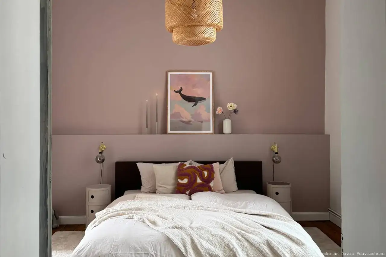 Sanfte Rosé-Wandfarbe hinter Bett mit Walbild, Kerzen und Blumen. Gemütliches Schlafzimmer mit Korb-Lampe.