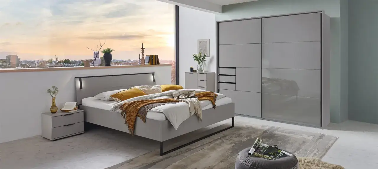 Modernes Schlafzimmer mit grauem Bett, Nachttischen und einem großen Kleiderschrank. Ein Fenster gibt den Blick auf eine Stadt bei Sonnenuntergang frei.