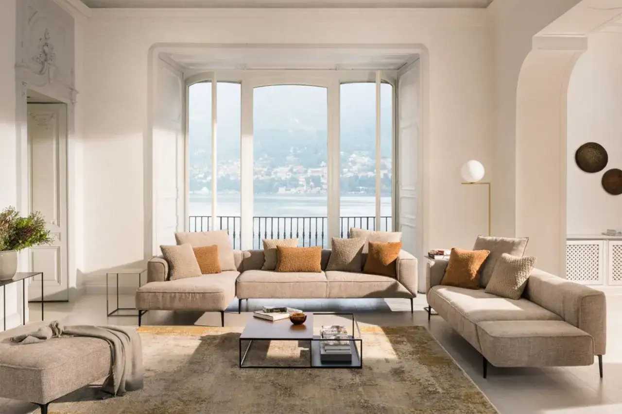 Ein Wohnzimmer, das zeigt, wie die Farbe Beige aussieht: Sofas, ein Teppich und ein Hocker in verschiedenen Beigetönen, akzentuiert durch orangefarbene Kissen.