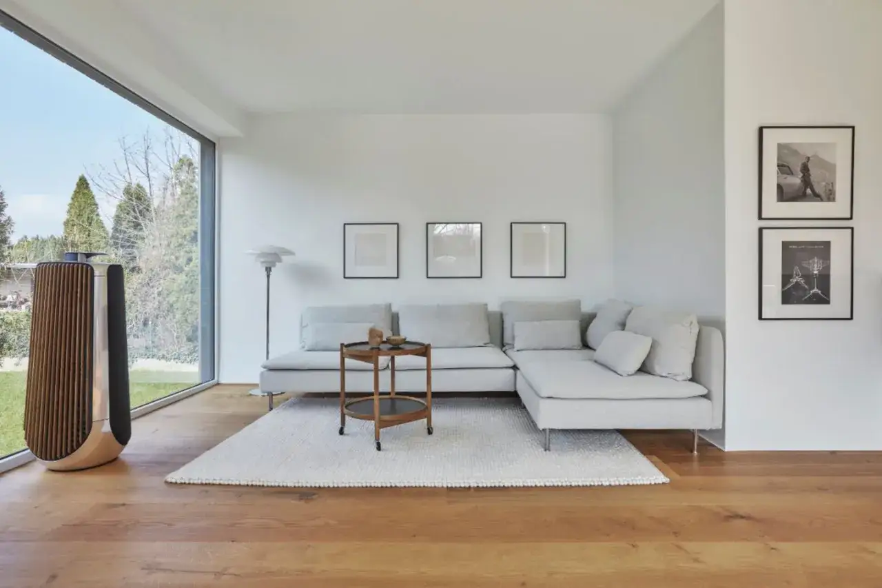 Ein Wohnzimmer in extrem minimalistischem Stil mit einem großen Fenster, einem hellgrauen Ecksofa, einem Beistelltisch und einem Lautsprecher.