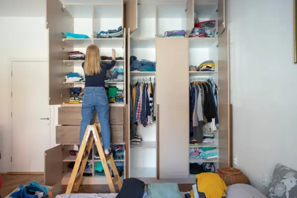 Mädchen auf Trittleiter sortiert Kleidung im offenen Kleiderschrank. So miste ich richtig aus: Kleidung wird auf Regalen und Kleiderstangen verstaut.
