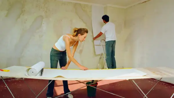 Ein Paar tapeziert eine gestrichene Wand. Die Frau misst Tapete auf einem Tisch, während der Mann sie an der Wand anbringt.