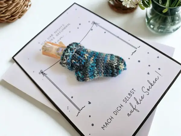 Gestrickte Socke mit Geld als Geschenk. Die Karte fragt "wie viele Socken braucht man?" und ermutigt, sich selbst auf die Socken zu machen.