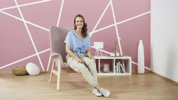 Frau sitzt in einem Sessel vor einer Wand mit geometrischem **Wanddesign streichen**. Ein Beistelltisch mit Deko steht daneben.